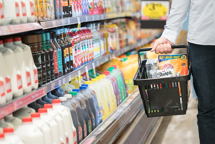 Shopper in grocery store holding full hand basket looking at cold drink aisle