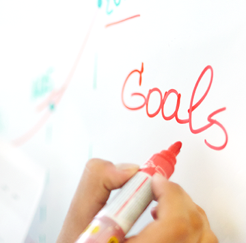 Person writing the word "Goals" on a white board with red pen